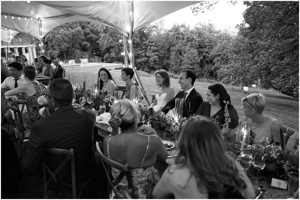 A bride and groom sit at a reception table listening to a wedding toast during an outdoor wedding in Iowa.