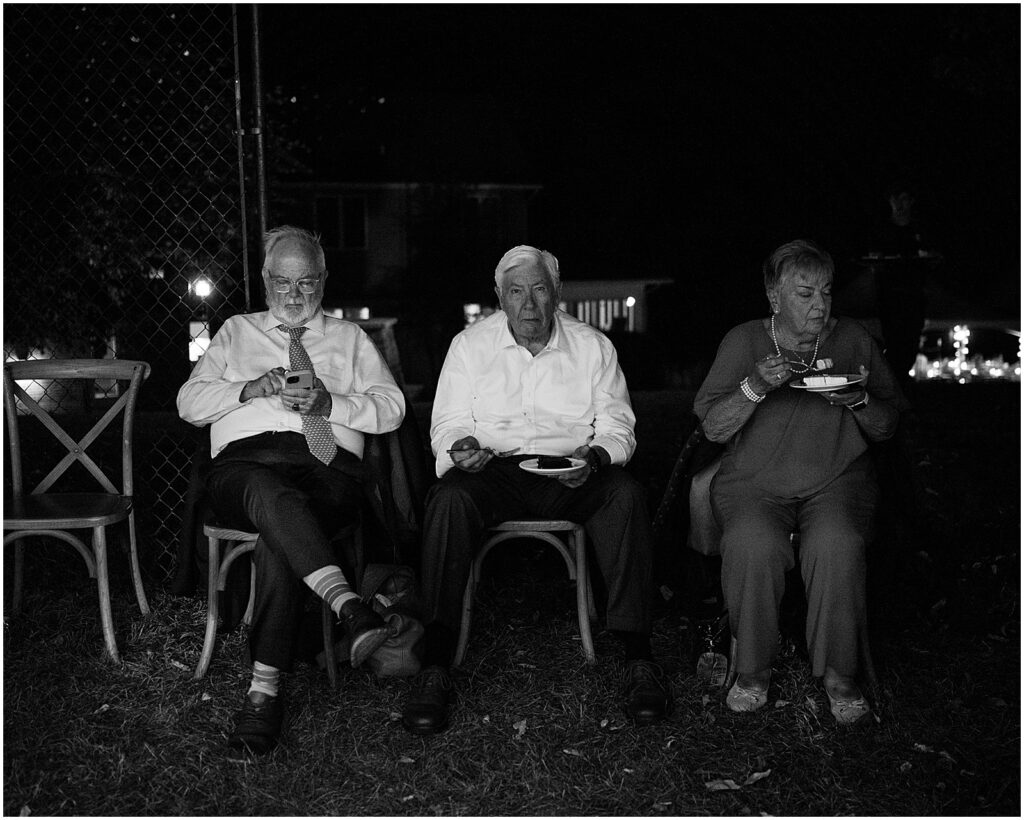 Elderly wedding guests sit on the edge of a dance floor eating cake.