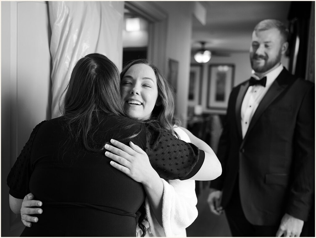 A bride hugs a family member while a groom smiles.