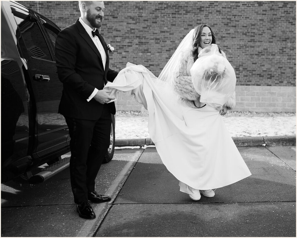 A groom carries a bride's train in a parking lot.