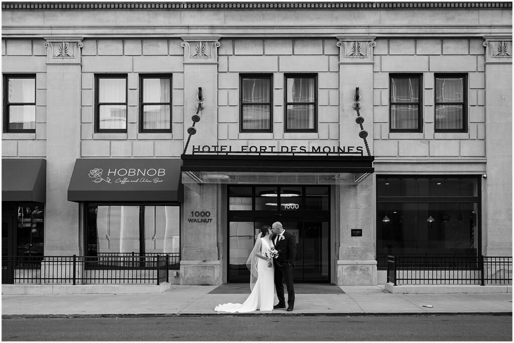 A bride and groom kiss on the sidewalk in front of Hotel Fort Des Moines.