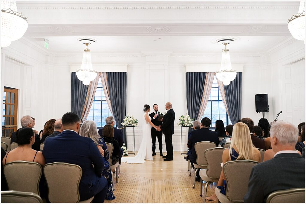 A bride and groom hold hands during a wedding ceremony in a hotel ballroom.