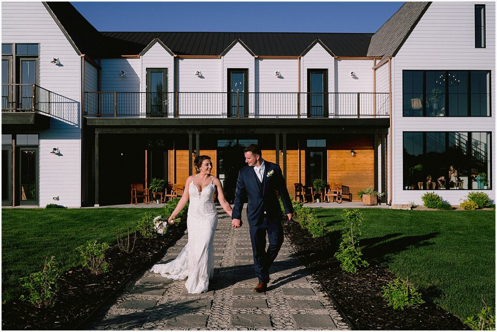 A bride and groom hold hands and walk down a path outside an Eastern Iowa wedding venue.