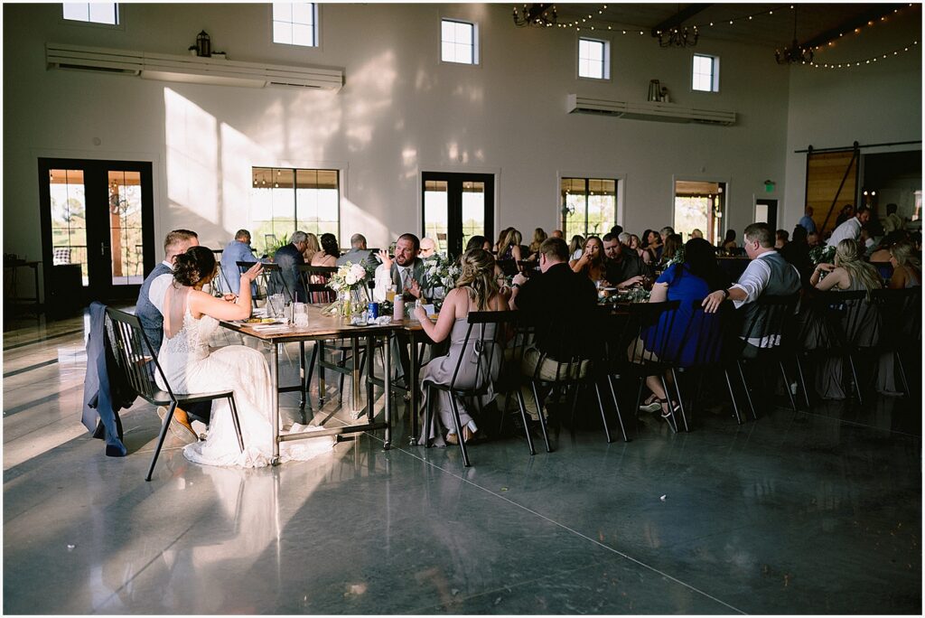 Wedding guests sit at long reception tables in a sunny wedding venue in Iowa.